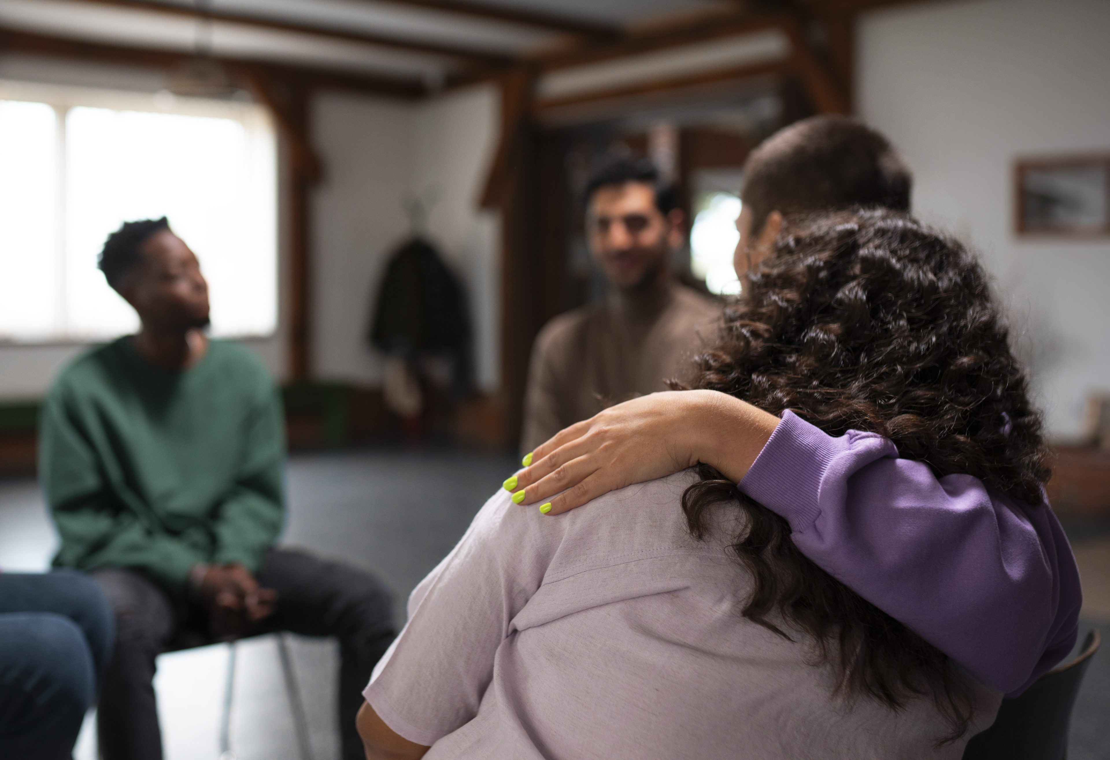 A support group meeting in a cozy room. A person in purple comforting another with a hand on the shoulder. Two others sit across, engaged and attentive.