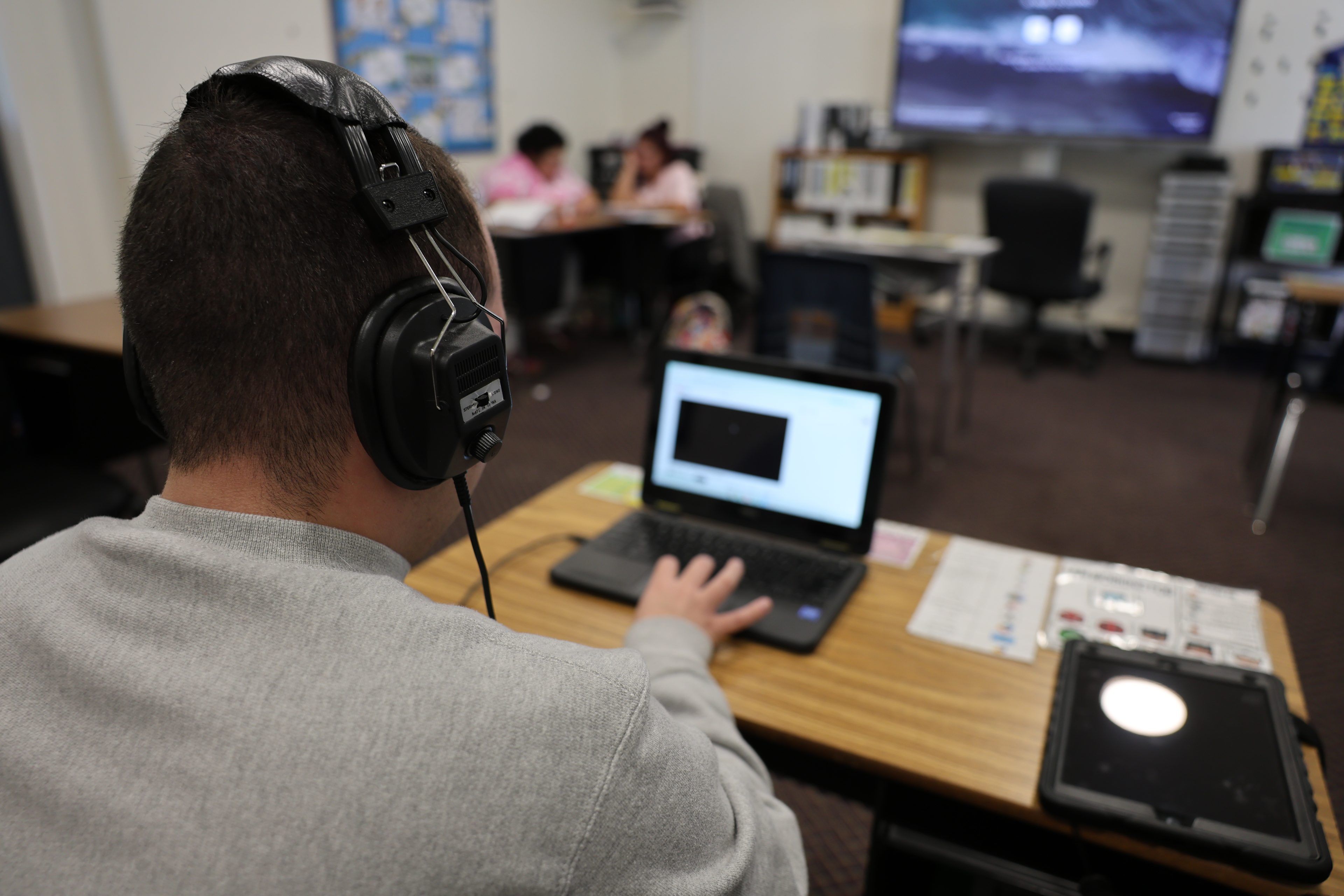 A person wearing headphones works on a laptop at a desk in a classroom setting, with other people and a large screen visible in the blurred background.