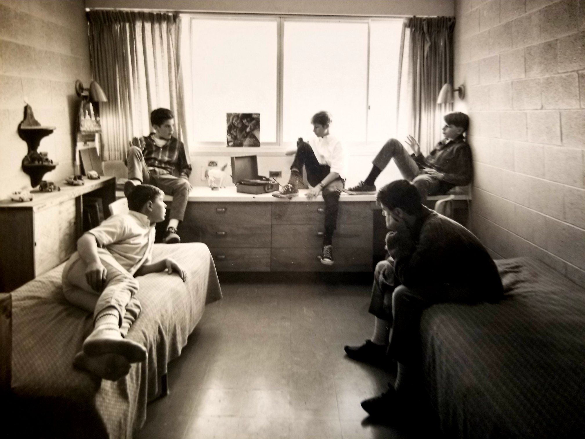 Five young men relax in a sunlit dorm room. Two sit on beds, three perch by a window. The scene feels casual and timeless, with subdued tones.