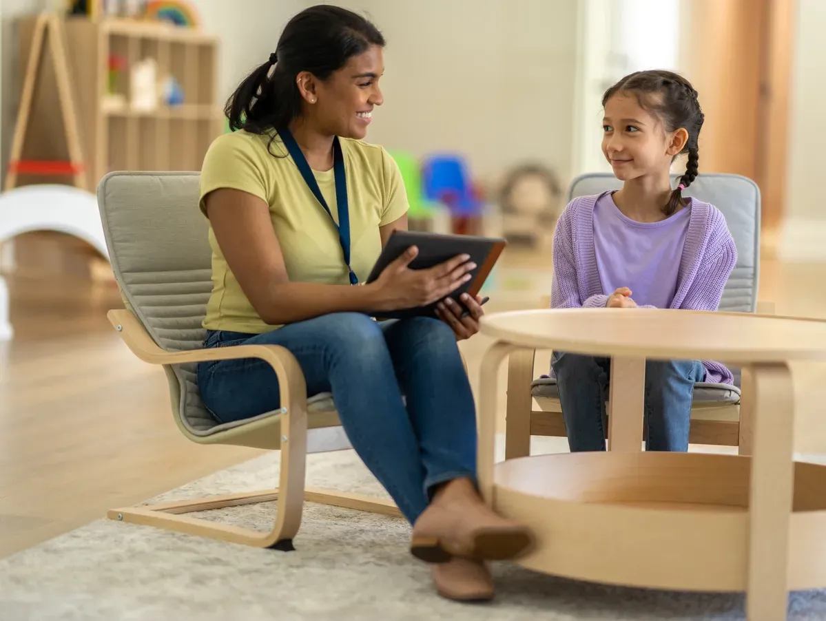A woman in casual clothes sits on a chair, holding a tablet, smiling at a young girl on a sofa. They are in a cozy room, conveying a warm, friendly atmosphere.