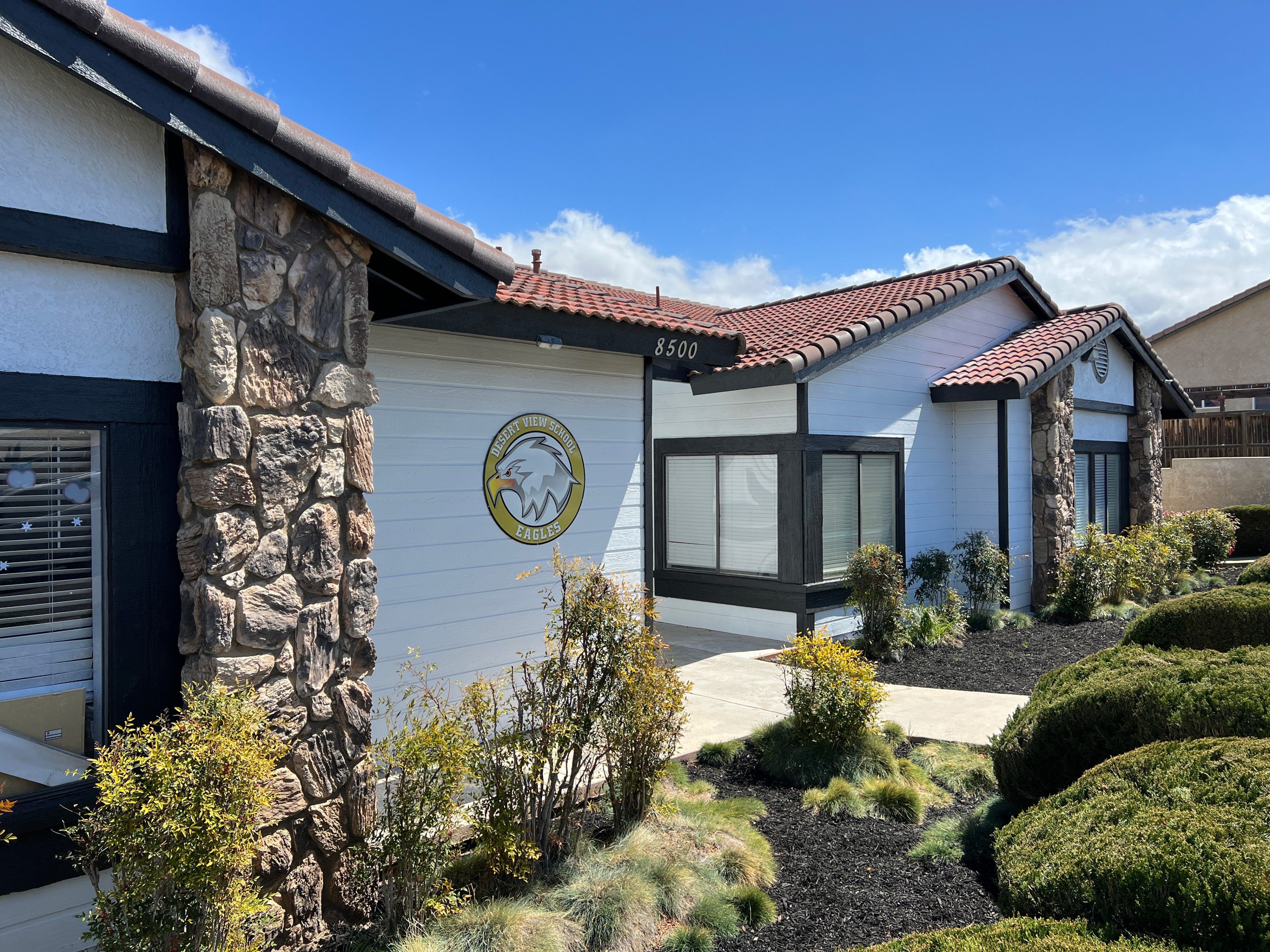 The image shows a sunny exterior of a modern building with stone and white walls, red-tiled roof, and landscaped greenery, under a clear blue sky.