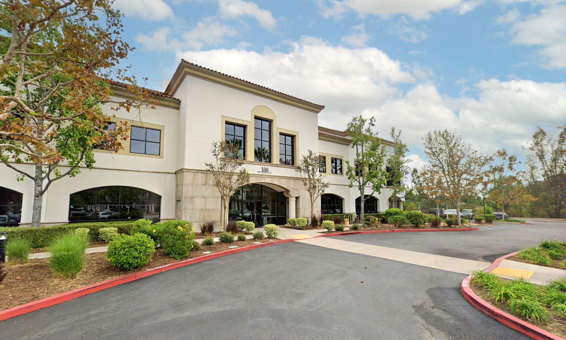 A modern two-story office building with white walls and large windows, surrounded by manicured greenery and a clear blue sky, conveying a professional ambiance.